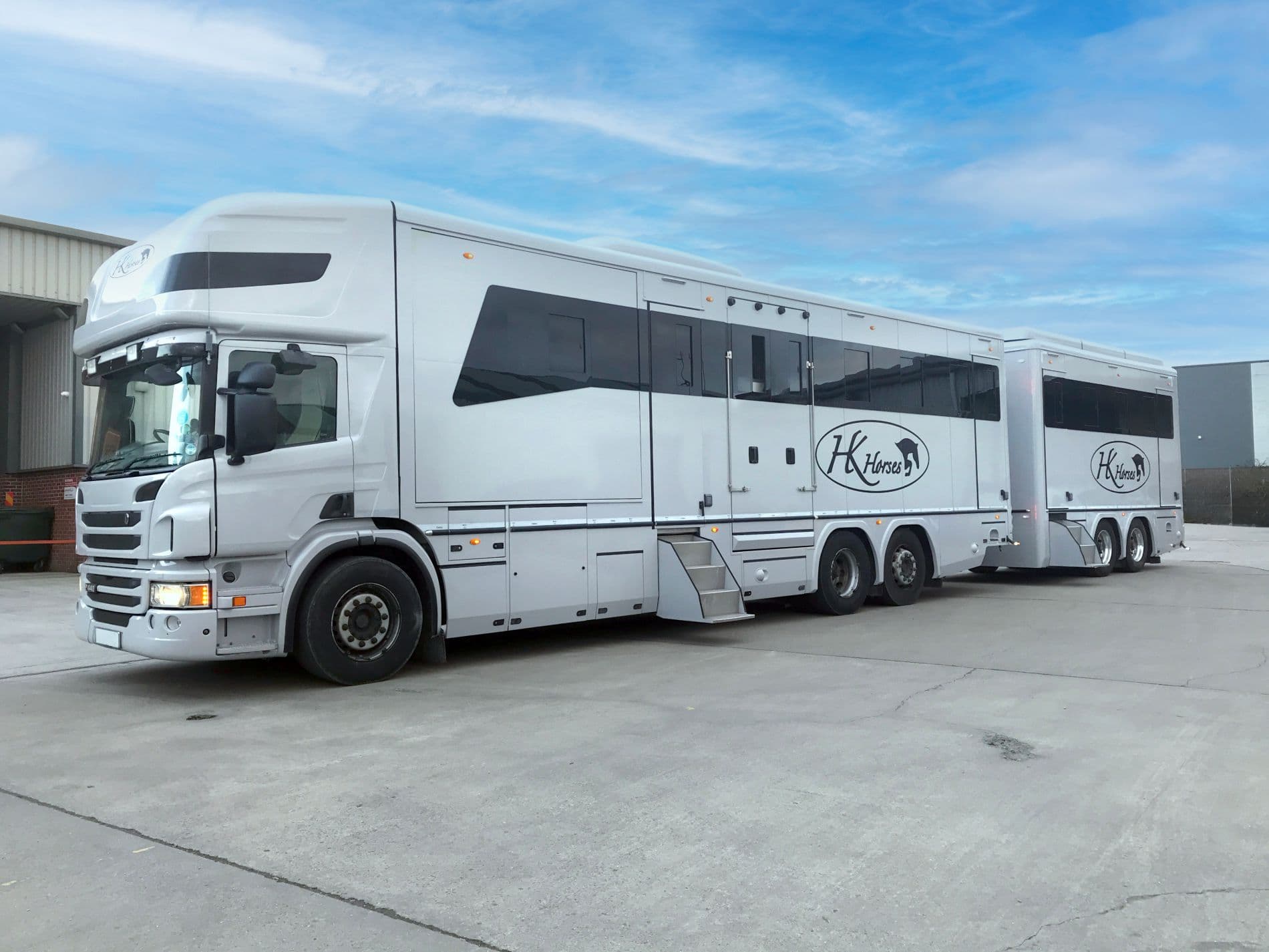 A white Scania HGV horsebox with a custom coach-built body, featuring integrated living quarters and horse transport area, parked on a concrete lot under a blue sky.